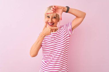 Orta yaş kadın çizgili t-shirt izole pembe arka plan üzerinde ayakta giyen mutlu yüz ile elleri ve parmakları ile çerçeve yapma gülümseyerek. Yaratıcılık ve fotoğrafçılık konsepti.