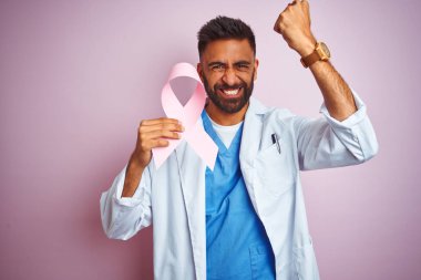 Genç hint doktor adam izole pembe arka plan üzerinde duran kanser kurdele tutan rahatsız ve öfke ile bağırarak sinirli, deli ve yükseltilmiş el ile bağırıyor, öfke kavramı