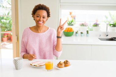 Genç afro-amerikan kadın evde sabah breaksfast yeme mutlu yüzü ile gülümseyerek zafer işareti yapıyor kameraya göz kırparak. İki numara.