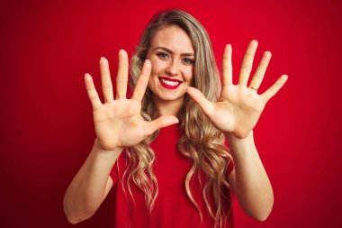 Genç güzel kadın kırmızı izole arka plan üzerinde duran temel t-shirt giyen gösteren ve emin ve mutlu gülümserken parmakları on numara ile işaret.