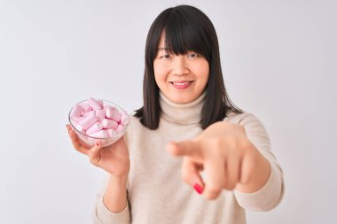 Kamera ve size parmak ile işaret izole beyaz arka plan üzerinde marshmallows ile kase tutan güzel Çinli kadın, el işareti, önden olumlu ve kendinden emin jest