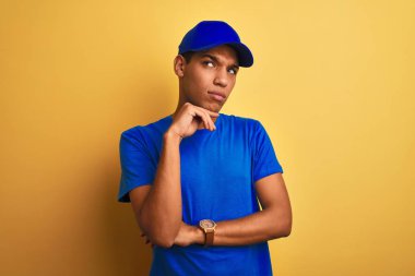 Genç yakışıklı arap teslimatadam çene üzerinde el ile izole sarı arka plan üzerinde duran soru, dalgın ifade düşünme. Düşünceli bir yüzle gülümsüyor. Şüphe kavramı.