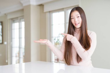 Beyaz masada rahat kazak giyen güzel Asyalı kadın şaşırttı ve el ile sunarken kameraya gülümseyerek ve parmak ile işaret.