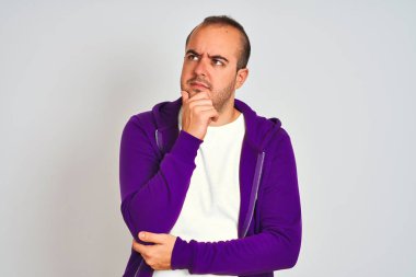 Mor sweatshirt giyen genç adam çenesi üzerinde el ile izole beyaz arka plan üzerinde duran soru, dalgın ifade düşünüyor. Düşünceli bir yüzle gülümsüyor. Şüphe kavramı.