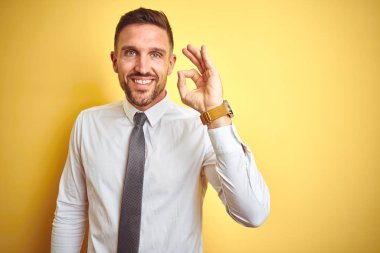 Genç yakışıklı iş adamı sarı izole arka plan üzerinde zarif beyaz gömlek giyen el ve parmakları ile ok işareti yapıyor olumlu gülümseyerek. Başarılı ifade.