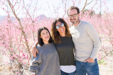 Üç gülümseyen güzel ailesi neşeli ve sarılma şeftali bahçesinde bahar güneşli gün zevk pembe yaprakları ile