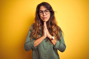 Yelllow izole arka plan üzerinde yeşil gömlek ve gözlük giyen genç güzel kadın birlikte emin gülümseyerek af isteyen elleri ile dua.