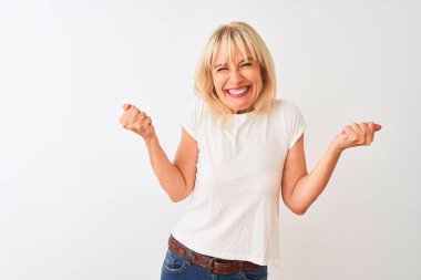 Orta yaş kadın izole beyaz arka plan üzerinde duran rahat t-shirt giyen çok mutlu ve heyecanlı kolları kaldırdı ile kazanan jest yapıyor, gülümseyerek ve başarı için çığlık. Kutlama konsepti.
