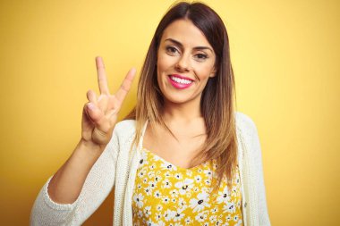 Genç güzel kadın sarı izole arka plan üzerinde duran ceket giyen gösteren ve emin ve mutlu gülümserken parmakları ile iki numara işaret.