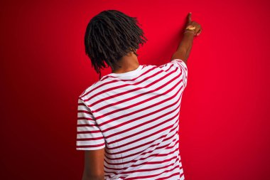 İzole kırmızı arka plan üzerinde duran çizgili t-shirt giyen dreadlocks giyen genç afro adam parmak el ile ileriye işaret geriye doğru poz