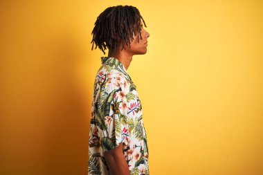Yan bakarak izole sarı arka plan üzerinde yazlık gömlek giyen tatilde dreadlocks ile Afro adam, kendine güvenen gülümseme ile doğal yüzü ile profil poz rahatla.