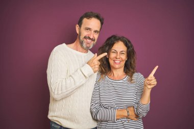 Güzel orta yaş çift yüzünde büyük bir gülümseme ile izole mor arka plan üzerinde kış kazak giyen, kameraya bakarak yan el ve parmak ile işaret.
