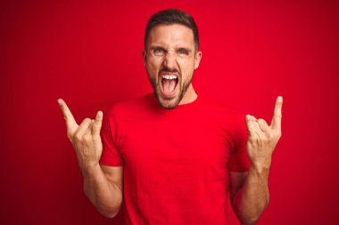 Genç yakışıklı adam kırmızı izole arka plan üzerinde rahat t-shirt giyen elleri ile rock sembolü yapıyor deli ifade ile bağırarak. Müzik yıldızı. Ağır müzik konsepti.