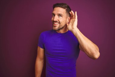 Lila izole arka plan üzerinde rahat mor t-shirt giyen genç adam söylenti veya dedikodu bir işitme dinlerken kulak üzerinde el ile gülümseyerek. Sağırlık kavramı.