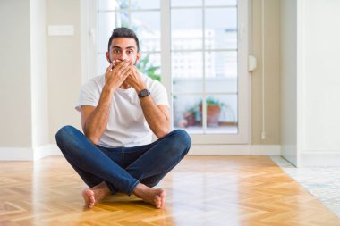 Evde yerde oturan rahat tişört giyen yakışıklı İspanyol adam hata yaptığı için elleriyle ağzını kapatarak şok etti. Gizli kavram.