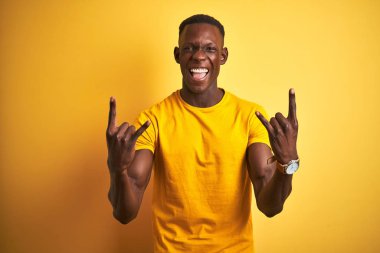 Genç afro-Amerikan adam rahat t-shirt izole sarı arka plan üzerinde duran deli ifade elleri ile rock sembolü yapıyor bağırarak. Müzik yıldızı. Ağır müzik konsepti.