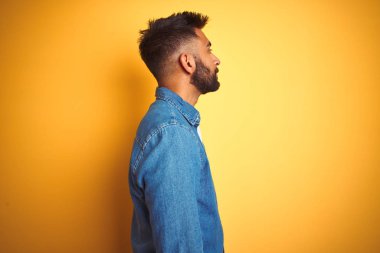 Denim gömlek giyen genç hintli adam yan bakarak izole sarı arka plan üzerinde duran, kendine güvenen gülümseme ile doğal yüzü ile profil poz rahatlayın.