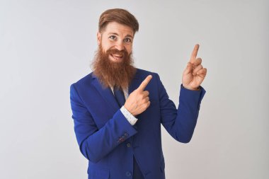 Genç kızıl saçlı İrlandalı işadamı izole beyaz arka plan üzerinde duran takım elbise giyen gülümseyerek ve yan iki el ve parmakları ile işaret kamera bakarak.