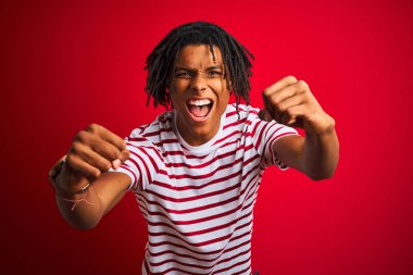 Çizgili t-shirt giyen dreadlocks giyen genç afro adam izole kırmızı arka plan üzerinde duran kızgın ve deli yükselterek yumruklar sinirli ve öfkeli öfke ile bağırırken. Öfke ve agresif kavram.