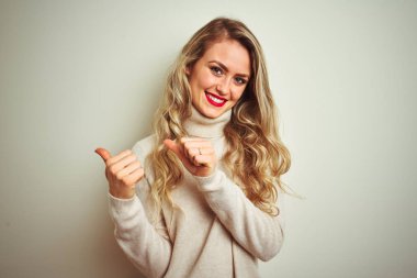 İzole beyaz arka plan üzerinde kış boğazlı kazak giyen güzel kadın el ve başparmak yukarı arka işaret, kendinden emin gülümseyerek