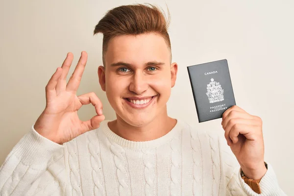 Young handsome man holding Canada Canadian passport over isolated white background doing ok sign with fingers, excellent symbol