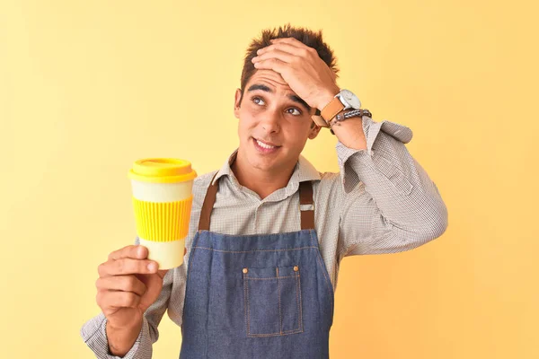 Young handsome barista man wearing apron holding coffee over isolated yellow background stressed with hand on head, shocked with shame and surprise face, angry and frustrated. Fear and upset for mistake.