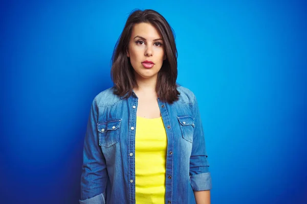 Young beautiful brunette woman wearing casual blue denim jacket over blue isolated background with serious expression on face. Simple and natural looking at the camera.