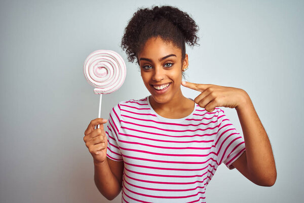 Африканская американская подросток женщина ест красочные конфеты на изолированном белом фоне очень счастливо указывая рукой и пальцем
