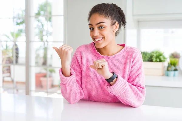 Beautiful african american woman with afro hair wearing casual pink sweater Pointing to the back behind with hand and thumbs up, smiling confident