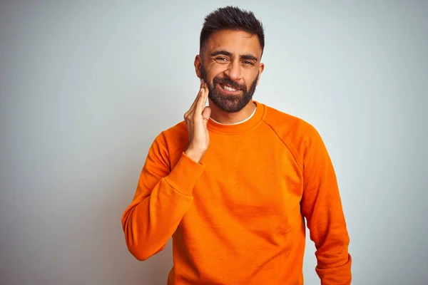 Young indian man wearing orange sweater over isolated white background touching mouth with hand with painful expression because of toothache or dental illness on teeth. Dentist concept.