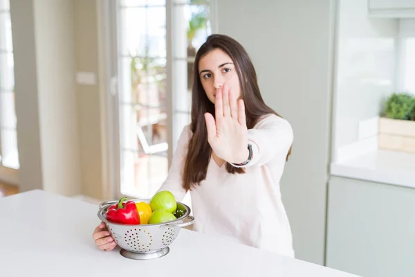 Beautiful young woman using colander to wash and clean vegatables with open hand doing stop sign with serious and confident expression, defense gesture