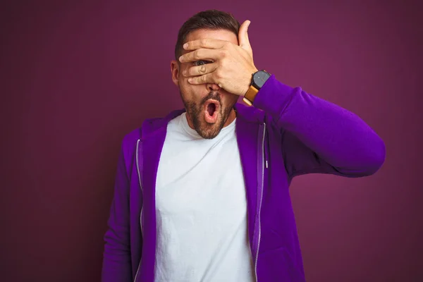 紫色の孤立した背景の上にカジュアルなスポーツスウェットシャツを着た若いフィットネスマンは 恥ずかしい表情で指を通して顔と目を覆うショックで覗く — ストック写真