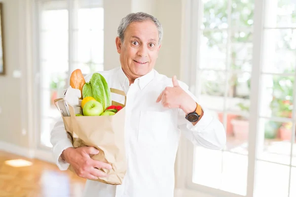 Handsome senior man holding a paper bag of fresh groceries from the supermarket with surprise face pointing finger to himself