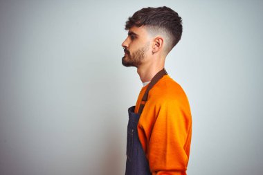 Yan bakarak izole beyaz arka plan üzerinde duran önlük giyen dövme giyen genç esnaf adam, kendine güvenen gülümseme ile doğal yüzü ile profil poz dinlenmek.