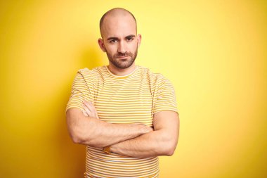 Sakallı genç kel adam sarı izole arka plan şüpheci ve sinir üzerinde rahat çizgili t-shirt giyiyor, çapraz kolları ile yüzünde onaylamayan ifade. Negatif kişi.