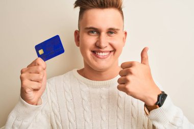 Genç yakışıklı adam izole beyaz arka plan üzerinde kredi kartı tutan büyük bir gülümseme ok işareti yapıyor mutlu, parmakları ile başparmak, mükemmel işareti