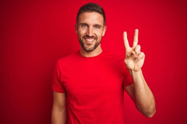 Genç yakışıklı adam kırmızı izole arka plan üzerinde rahat t-shirt giyen kameraya bakarak gülümseyerek zafer işareti yapıyor parmakları gösteren. İki numara.