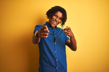 Rahat kot gömlek giyen dreadlocks ile Afro adam mutlu ve komik yüzü ile kameraya parmakları gösteren izole sarı arka plan üzerinde duran. İyi enerji ve titreşimler.