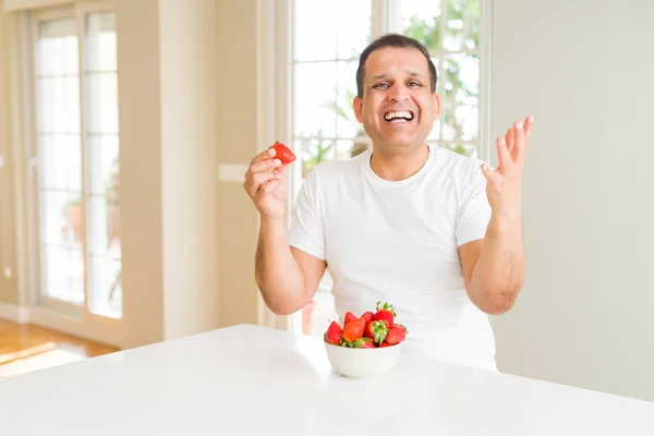 Middle age man eating strawberries at home very happy and excited, winner expression celebrating victory screaming with big smile and raised hands