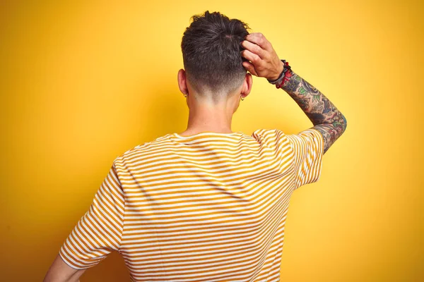 孤立した黄色の背景の上に立つストライプのTシャツを着たタトゥーを持つ若い男は 頭の上に手で疑いを考える後方に — ストック写真