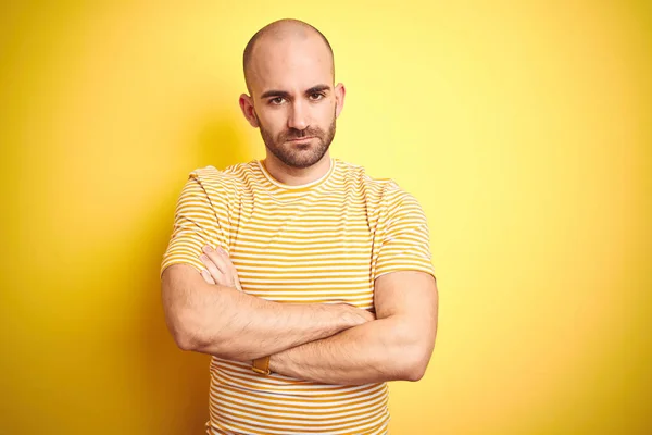 Young bald man with beard wearing casual striped t-shirt over yellow isolated background skeptic and nervous, disapproving expression on face with crossed arms. Negative person.