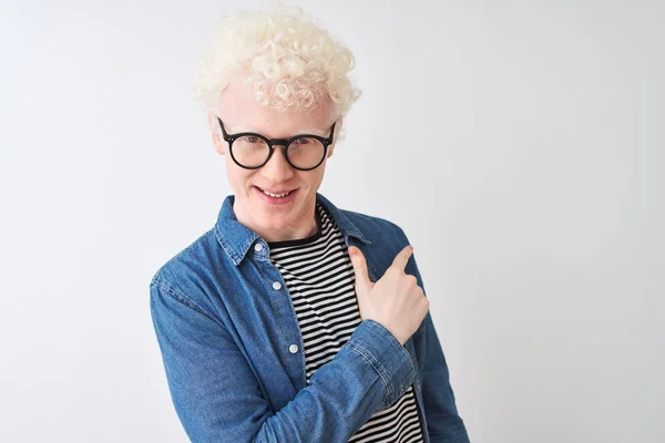 Young albino blond man wearing denim shirt and glasses over isolated white background cheerful with a smile on face pointing with hand and finger up to the side with happy and natural expression