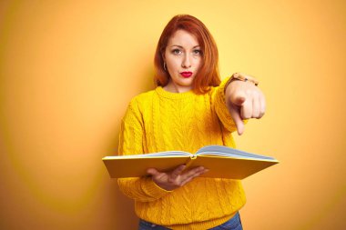 Genç kızıl saçlı öğrenci kadın kamera ve size parmak ile işaret sarı izole arka plan üzerinde duran kitap okuma, el işareti, önden olumlu ve kendinden emin jest