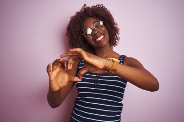 Genç Afrikalı afro kadın izole pembe arka plan üzerinde çizgili t-shirt gözlük giyen aşk kalp sembolü ve elleriyle şekil gösteren gülümseyerek. Romantik kavram.