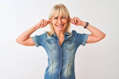 Orta yaş kadın yüksek sesle müzik sesi için rahatsız ifade ile parmakları ile kulakları kapsayan izole beyaz arka plan üzerinde duran rahat kot gömlek giyiyor. Sağır kavramı.