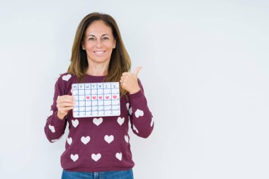 Orta yaş kadın izole arka plan üzerinde adet takvimi tutan işaret ve mutlu yüz gülümseyerek yan başparmak ile gösteren