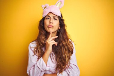 Pijama ve uyku maskesi takan genç kadın sarı izole arka plan üzerinde duran çene parmak ile şüphe hakkında konsantre ve merak bakarak