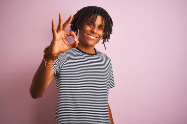 Dreadlocks lacivert çizgili t-shirt izole pembe arka plan üzerinde duran giyen Afro adam el ve parmaklar ile ok işareti yapıyor olumlu gülümseyerek. Başarılı ifade.