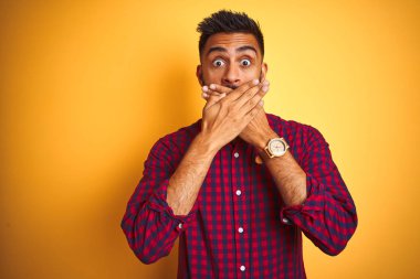 Izole sarı arka plan üzerinde duran rahat gömlek giyen genç Hintli adam hata için elleri ile ağız kapsayan şok. Gizli kavram.