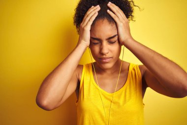 Izole sarı arka plan üzerinde kulaklık kullanarak müzik dinleyen Afro-Amerikan kadın baş ağrısı umutsuz muzdarip ve stresli çünkü ağrı ve migren. Eller baş üstünde.